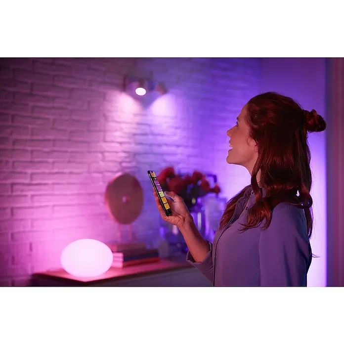 Philips Hue Smart-LED-Lampen Set White & Color - E27, Dimmbar, Warmweiß, 1.600 lm, 13,5 W, 1 Stk. - Frau benutzt eine App, um die farbigen intelligenten Philips Hue-Leuchten in ihrem Haus zu steuern und anzupassen.