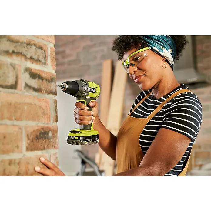 Ryobi ONE+ Taladro atornillador percutor de batería + caja herramientas y accesorios R18PD3-220TAH - 18 V, 2 baterías, 2 Ah, 50 Nm - Una mujer usa un taladro inalámbrico Ryobi para perforar un agujero en una pared de ladrillo mientras hace mejoras en el hogar.