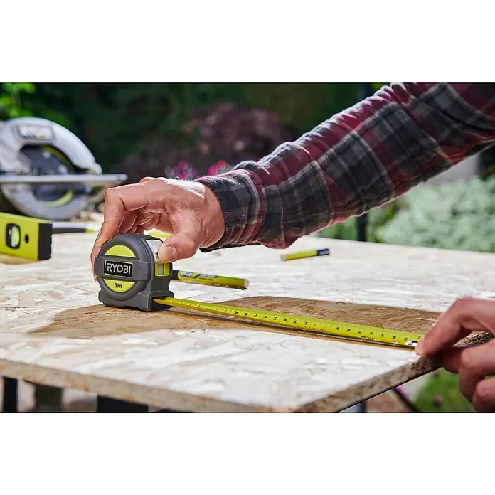 Ryobi ONE+ Taladro atornillador percutor de batería + caja herramientas y accesorios R18PD3-220TAH - 18 V, 2 baterías, 2 Ah, 50 Nm - Primer plano de una cinta métrica Ryobi que se utiliza sobre una superficie de madera con herramientas al fondo.