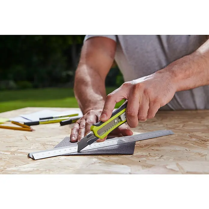 Ryobi ONE+ Taladro atornillador percutor de batería + caja herramientas y accesorios R18PD3-220TAH - 18 V, 2 baterías, 2 Ah, 50 Nm - El hombre usa un cúter para cortar un trozo de material a lo largo de una regla de metal para cortes precisos.