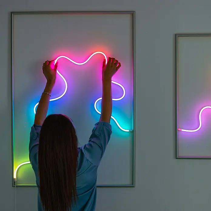 Twinkly LED-Lichtschlauch Flex - Länge: 2 m, Lichtfarbe: RGB, 15 W - Frau arrangiert eine Neonkunstinstallation mit bunten Lichtern in einem Rahmen an einer Wand.