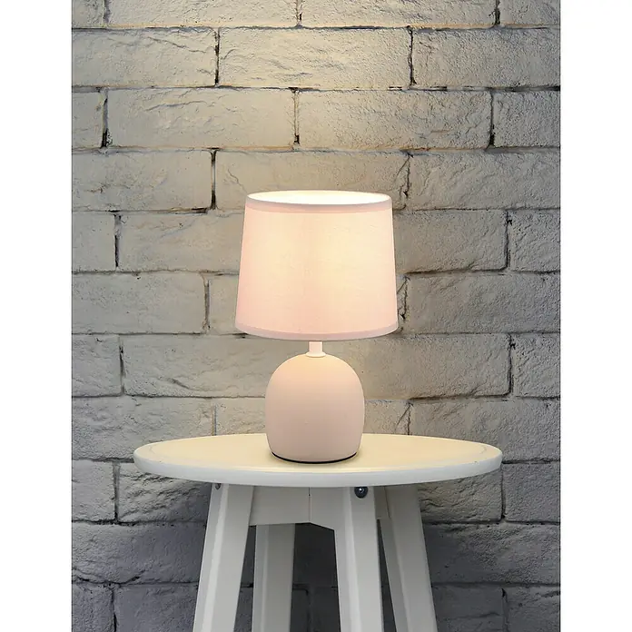 Trio Lighting Lámpara de sobremesa redonda Malu - 40 W, Ø x Al: 16 cm x 26 mm, Beige, Natural, E14 - Primer plano de una elegante lámpara de mesa con una pantalla de color crema, sobre una mesa auxiliar blanca frente a una pared de ladrillo.