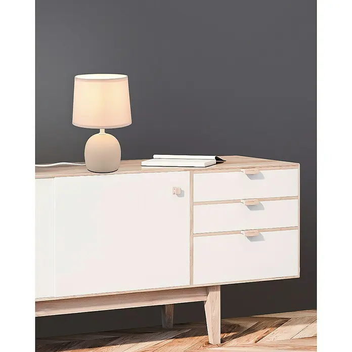Trio Lighting Lámpara de sobremesa redonda Malu - 40 W, Ø x Al: 16 cm x 26 mm, Beige, Natural, E14 - Una elegante lámpara de mesa y un libro sobre un aparador blanco con cajones y puerta, sobre una pared gris.