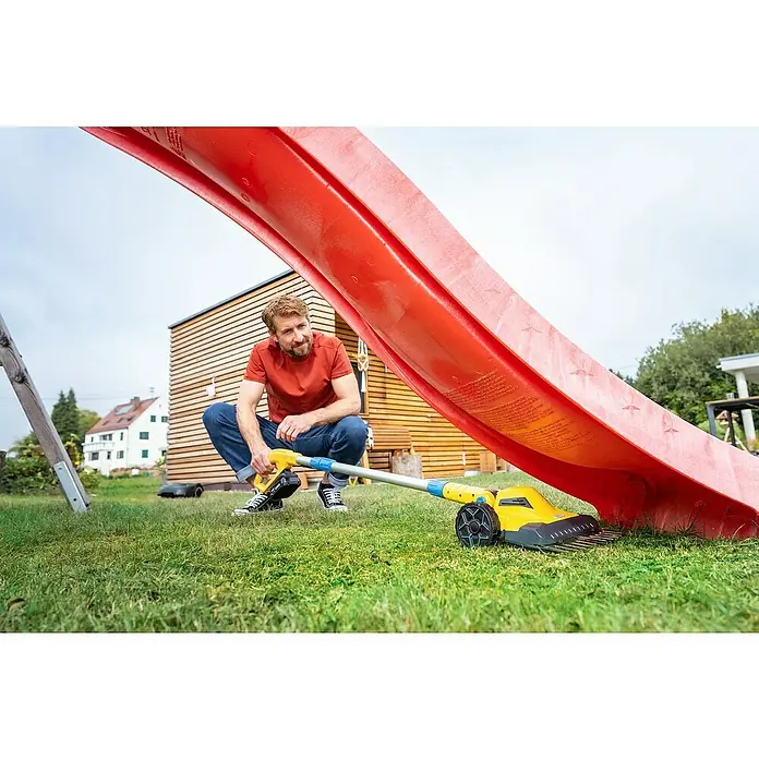 Gloria Power for All 18V Akku-Rasenmäher Grasschere FineCut 18V - 18 V, 1 Akku, Schnittbreite: 25 cm - Ein Mann benutzt einen gelben Rasenlüfter, um das Gras unter einer roten Kinderrutsche zu pflegen.