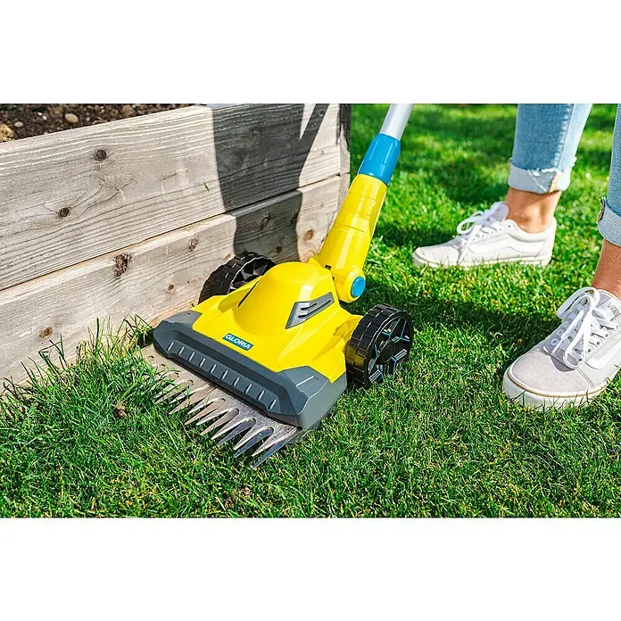 Gloria Power for All 18V Akku-Rasenmäher Grasschere FineCut 18V - 18 V, Ohne Akku, Schnittbreite: 25 cm - Das Bild zeigt einen gelben Gloria Rasenlüfter, der auf einem grünen Rasen neben einem erhöhten Blumenbeet steht.