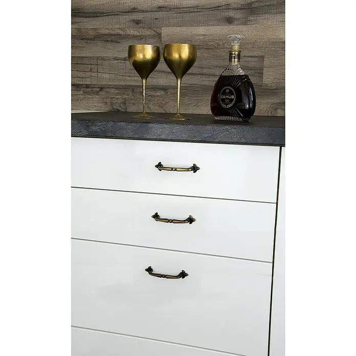 Siro Möbelgriff - Typ Möbelgriff: Bügel, L x B x H: 108 x 22 x 23 mm, Stahl, Braun - Nahaufnahme von weißen Schubladen mit goldenen Griffen, darüber zwei goldene Kelche und eine Flasche CAMUS XO Cognac.