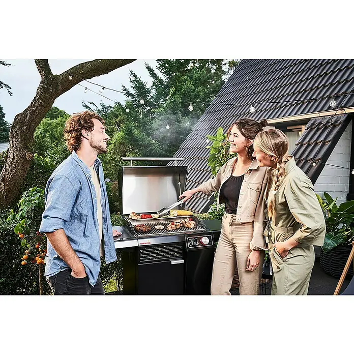 Char-Broil Elektrogrill Smart E - Grillfläche (B x T): 57 x 39 cm, 2,4 kW - Eine Gruppe von Freunden steht um einen modernen Grill herum, auf dem Fleisch und Gemüse zubereitet werden. Stimmungsvolle Gartenbeleuchtung sorgt für Atmosphäre.