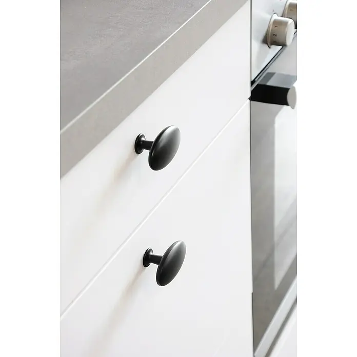 Siro Möbelknopf - Typ Möbelgriff: Knopf, L x B x H: 64 x 36 x 35 mm, Stahl, Schwarz - Schwarze ovale Möbelknöpfe an weißen Schubladen mit einer grauen Arbeitsplatte und einem Teil eines Ofens im Hintergrund.