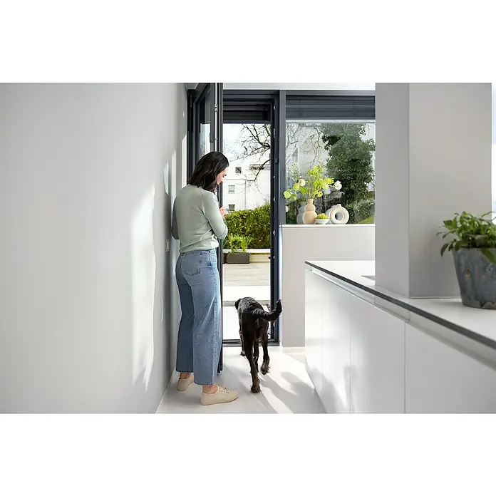 Bosch Smart Home Funk-Fensterkontakt II Plus - Anthrazit, Batteriebetrieben, 23 x 22 x 75 mm, 1 Stk. - Eine Frau öffnet eine moderne, rahmenlose Glastür mit einem eleganten schwarzen Hund, der neugierig nach draußen schaut.