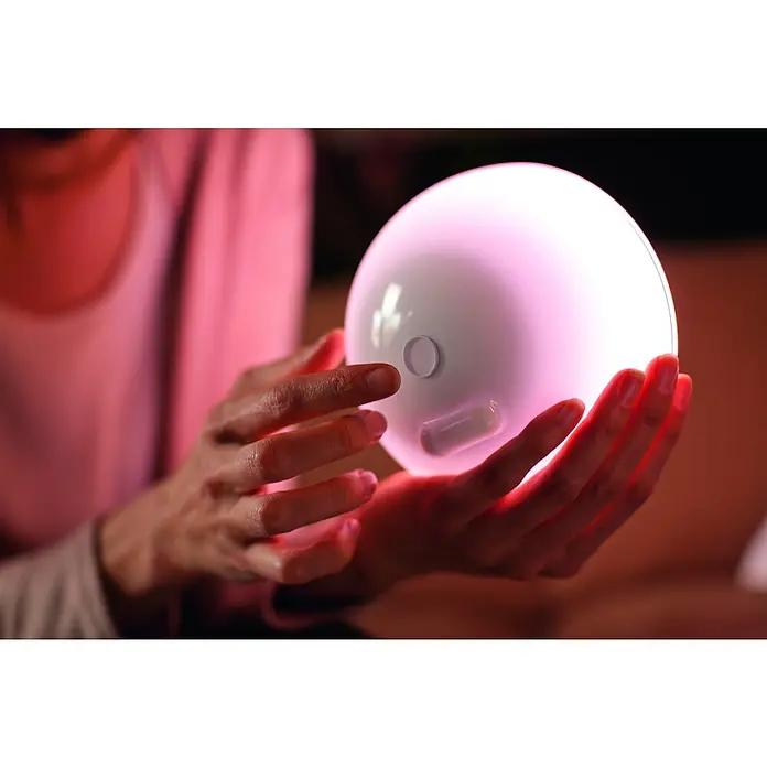 Philips Hue LED-Tischleuchte Go - 6 W, L x B x H: 15 x 15 x 7,9 cm, RGBW - Eine Nahaufnahme von Händen, die eine leuchtende, kugelförmige Philips Hue Lampe halten, die ein weiches, farbiges Licht ausstrahlt.