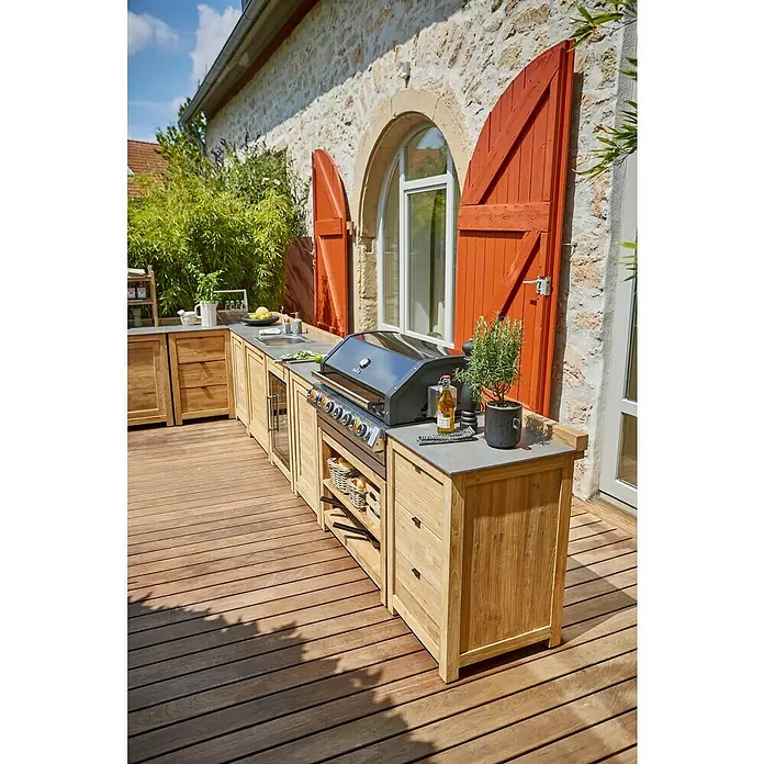 Sunfun Zoe Módulo de cocina exterior Armario con cajones - Marrón, An x Pr x Al: 58 x 63 x 92 cm, Madera de teca - Isla de cocina exterior con parrilla, fregadero y almacenamiento en una terraza de madera frente a una casa de piedra con contraventanas rojas.