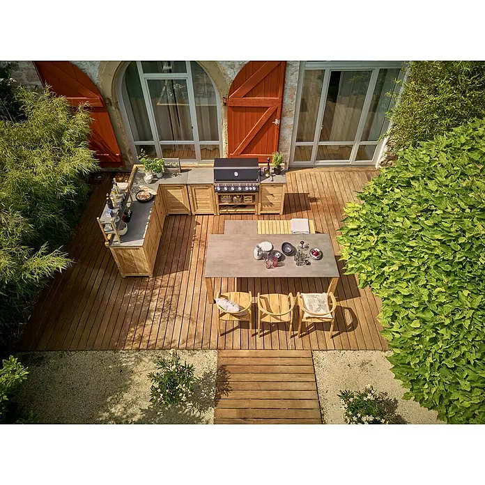 Sunfun Zoe Outdoorküchen-Modul Eckschrank - Braun, B x T x H: 120 x 120 x 92 cm, Teakholz - Eine Outdoor-Küche mit Grill und Esstisch auf einer Holzterrasse, umgeben von üppigem Grün. Ideal für sommerliche Grillabende im Freien.