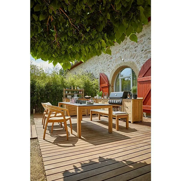 Sunfun Zoe Módulo de cocina exterior Armario con cajones - Marrón, An x Pr x Al: 58 x 63 x 92 cm, Madera de teca - Un acogedor espacio al aire libre con un moderno conjunto de mesa de comedor y barbacoa en una terraza de madera, rodeado de vegetación exuberante.