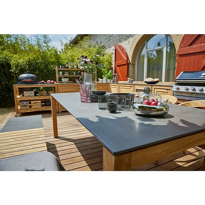 Sunfun Zoe Gartentisch - L x B x H: 220 x 100 x 77 cm, Keramik, Teak, Rechteckig, Naurbraun/Anthrazit - Ein großer Esstisch aus Holz mit grauer Oberfläche, gedeckt mit Geschirr und Dekorationen, steht im Freien auf einer Holzterrasse.