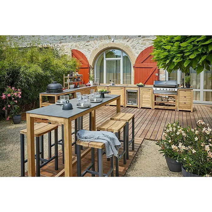 Sunfun Zoe Cocina exterior Armario bajo - Marrón, An x Pr x Al: 93 x 57,5 x 66 cm, Madera de teca - Un espacio exterior acogedor con una cocina de barbacoa, una mesa con taburetes y una exuberante vegetación para un ambiente relajante.