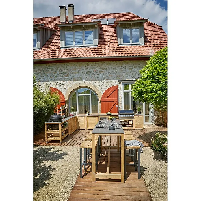 Sunfun Zoe Outdoorküchen-Modul Kamado - Braun, B x T x H: 120 x 71,5 x 89 cm, Teakholz - Eine Außenküche mit Grill und Esstisch auf einer Holzterrasse vor einem Haus mit roten Fensterläden.