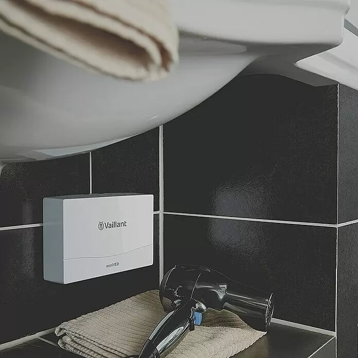 Vaillant Kleindurchlauferhitzer - 3,5 kW, Hydraulisch, Offen - Weißer Vaillant miniVED Durchlauferhitzer an einer dunkel gefliesten Wand unter einem Waschbecken, mit Föhn und Handtuch.