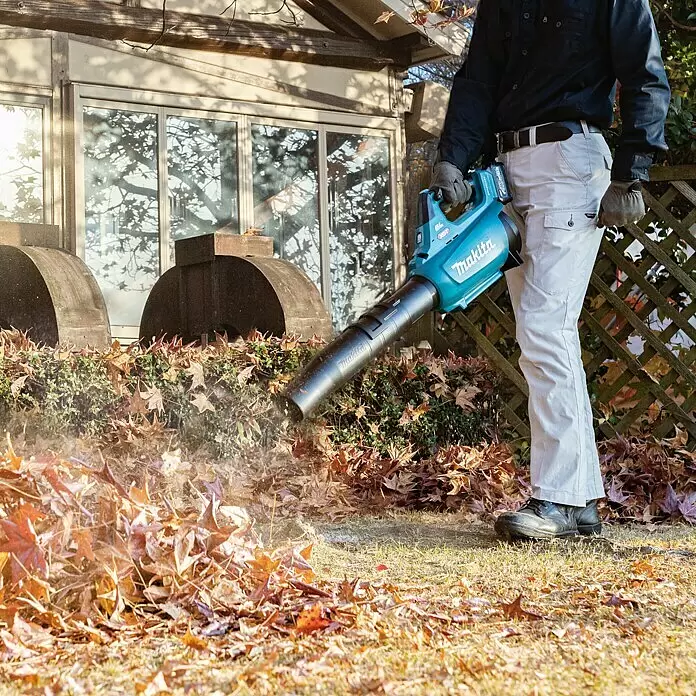 Makita Akku-Laubbläser UB001GZ - 40 V, Ohne Akku, Blasgeschwindigkeit: 53 m/s - Person bläst mit türkis-schwarzem Makita Laubbläser Herbstlaub in einem Garten weg.