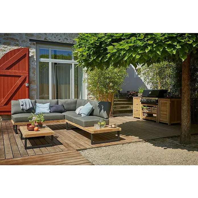 Sunfun Zoe Módulo de cocina exterior Armario con cajones - Marrón, An x Pr x Al: 58 x 63 x 92 cm, Madera de teca - Acogedora escena al aire libre con un sofá seccional gris, mesas de madera y una parrilla para barbacoa bajo un árbol sombrío.