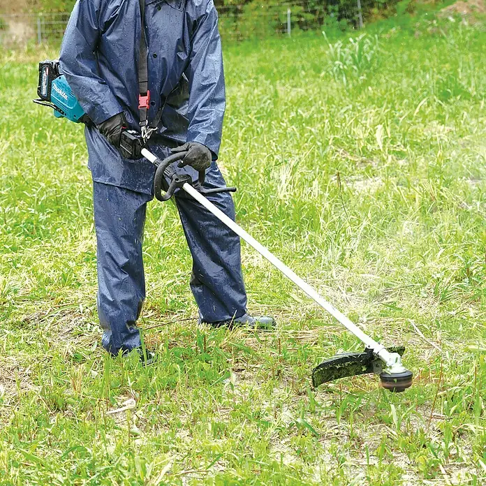 Makita XGT 40V max. Akku-Freischneider UR007GZ01 - 40 V, Ohne Akku, Schnittbreite Faden: 43 cm, Schnittbreite Messer: 25,5 cm - Person in Regenkleidung verwendet einen Makita Akku-Rasentrimmer im Gras. Der Trimmer ist mit einem Akku ausgestattet und hat einen Griff.