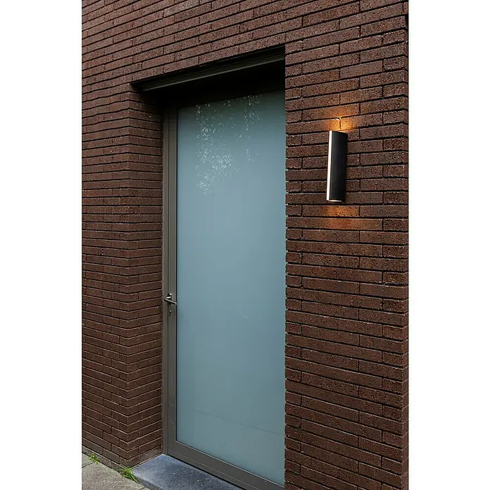 Lutec LED-Außenwandleuchte Leo - 14,5 W, 8,8 x 6,6 x 30 cm, Anthrazit, IP54 - Eine moderne schwarze Wandleuchte beleuchtet eine braune Backsteinmauer neben einer milchglasverkleideten Tür und erzeugt einen warmen Lichteffekt.