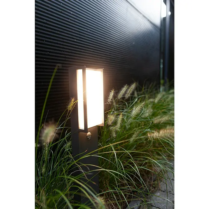 Lutec Qubo LED-WegeleuchteHöhe: 75 cm, Warmweiß - Die moderne Außenleuchte spendet warmes Licht und ist von dekorativem Gras umgeben. Elegantes Design für Ihren Garten.