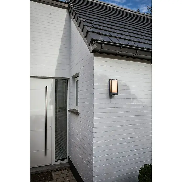 Lutec Qubo LED-Außenwandleuchte - 14 W, 10,6 x 11 x 27 cm, Anthrazit, IP54, Bewegungsmelder - Eine moderne rechteckige Wandleuchte mit warmem Licht schmückt eine weiße Backsteinfassade neben einem Hausfenster.