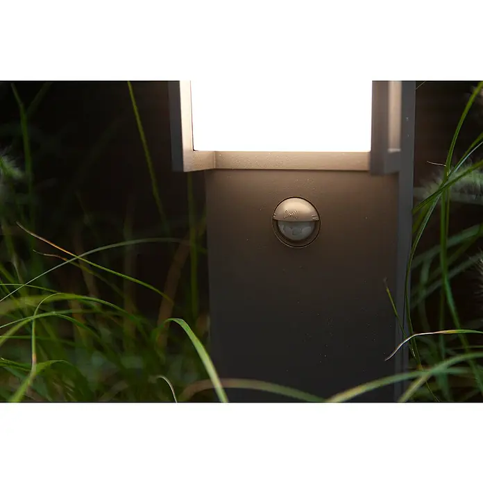 Lutec Qubo LED-WegeleuchteHöhe: 75 cm, Warmweiß - Nahaufnahme einer Außenleuchte mit Bewegungssensor, die ein warmes, einladendes Licht in der Nacht ausstrahlt. Die Lampe ist umgeben von grünen Gräsern.