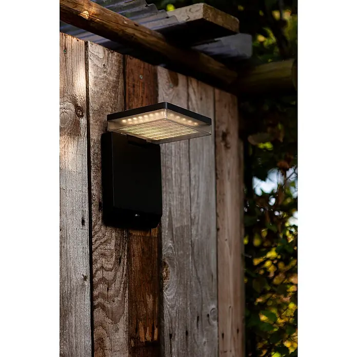 Lutec Solar-Außenwandleuchte Moze - 8 W, L x B x H: 19,5 x 14 x 15 cm, Schwarz matt, IP44, 1.000 lm - Eine moderne, schwarze LED-Wandleuchte mit Bewegungsmelder, die an einer Holzwand befestigt ist, strahlt helles Licht aus.