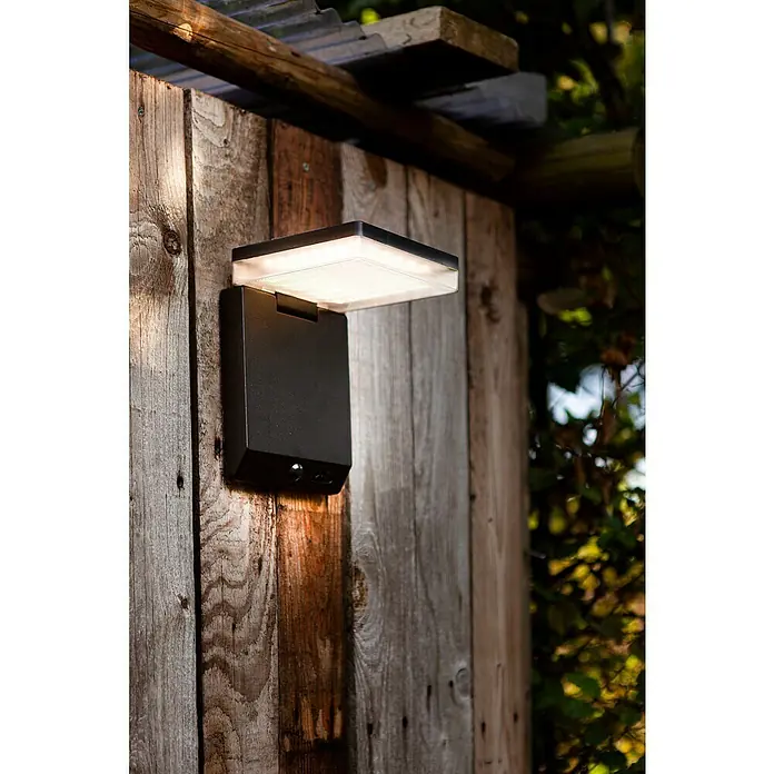 Lutec Solar-Außenwandleuchte Moze - 8 W, L x B x H: 19,5 x 14 x 15 cm, Schwarz matt, IP44, 1.000 lm - Eine moderne, schwarze Außenleuchte mit einem hellen, quadratischen Lichtelement, montiert an einer rustikalen Holzwand, die für Beleuchtung sorgt.