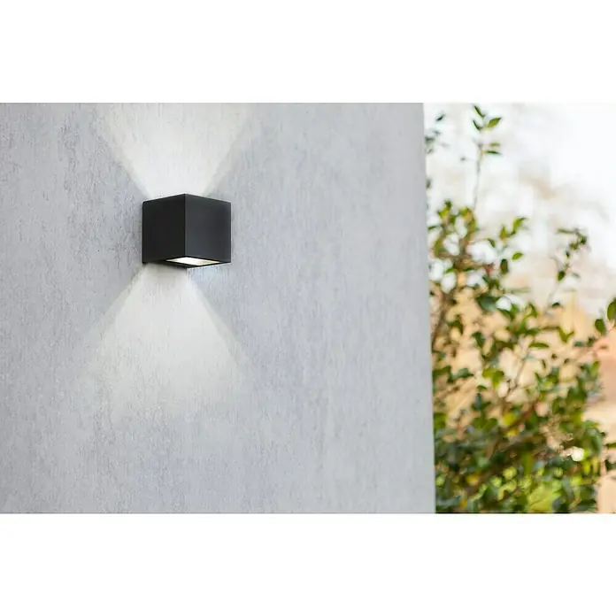 Lutec LED-Außenwandleuchte Gemini - 9 W, 8,8 x 8,5 x 8,6 cm, Schwarz matt, IP54 - Schwarze Würfel-Wandleuchte strahlt Licht nach oben und unten und erhellt eine strukturierte Wand mit Außenbepflanzung im Hintergrund.