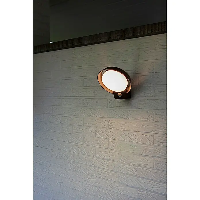 Lutec Polo LED-Sensor-Außenwandleuchte - 16,1 W, L x B x H: 18 x 16,4 x 16,2 cm, Anthrazit, Warmweiß - Nahaufnahme einer eleganten, runden Wandleuchte mit Bewegungssensor, die ein warmes, einladendes Licht auf eine strukturierte Backsteinmauer wirft.