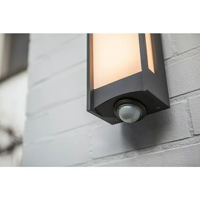 Lutec Qubo LED-Außenwandleuchte - 14 W, 10,6 x 11 x 27 cm, Anthrazit, IP54, Bewegungsmelder - Nahaufnahme einer beleuchteten Außenwandleuchte mit Bewegungssensor an einer weißen Ziegelwand.