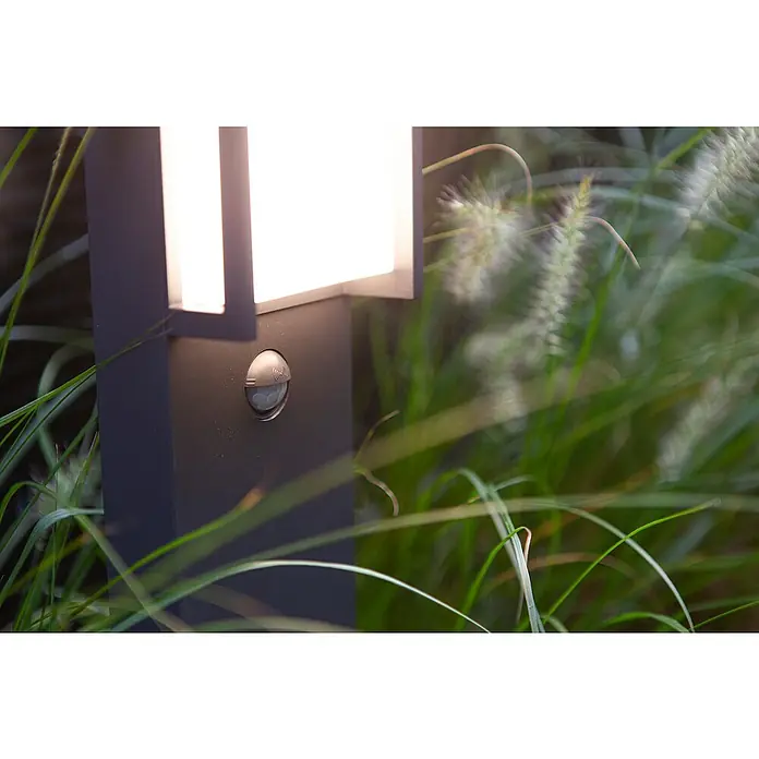 Lutec Qubo LED-WegeleuchteHöhe: 75 cm, Warmweiß - Nahaufnahme einer modernen Außenleuchte mit Bewegungssensor, eingebettet in grüne Pflanzen. Die Lampe strahlt helles Licht aus.