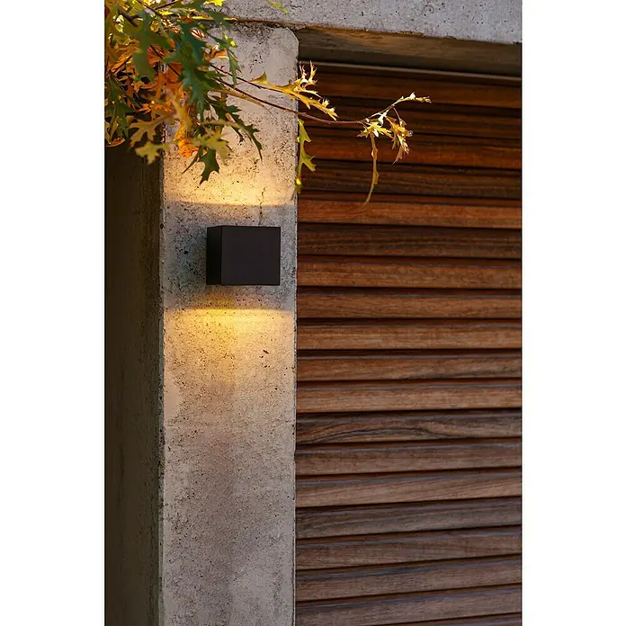 Lutec LED-Außenwandleuchte Gemini - 9 W, 8,8 x 8,5 x 8,6 cm, Schwarz matt, IP54 - Diese Nahaufnahme zeigt eine moderne, würfelförmige Außenwandleuchte, die ein warmes Licht auf eine Betonwand und eine Holzverkleidung wirft.