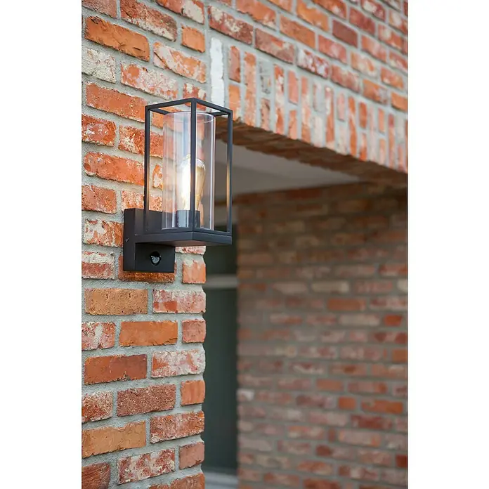 Lutec Solar-Außenwandleuchte FLAIR - 40 W, L x B x H: 17,2 x 12 x 31,3 cm, IP44, Nach oben gerichtet - Eine moderne schwarze Außenleuchte mit Bewegungsmelder an einer roten Backsteinmauer. Die Lampe hat eine klare, zylindrische Glasabdeckung.