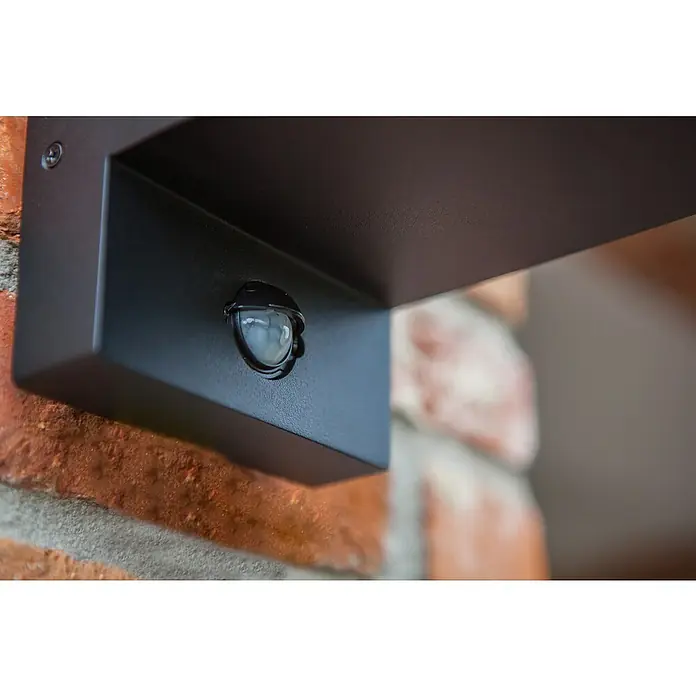 Lutec Solar-Außenwandleuchte FLAIR - 40 W, L x B x H: 17,2 x 12 x 31,3 cm, IP44, Nach oben gerichtet - Nahaufnahme einer schwarzen Außenleuchte mit Bewegungssensor an einer Ziegelwand, die moderne Beleuchtung und Sicherheit vereint.