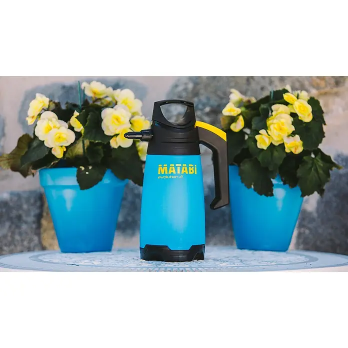 Matabi Sulfatadora Evolution 2 - Plástico - Primer plano de un pulverizador a presión azul Matabi Evolution 2 sobre una mesa, flanqueado por dos macetas azules con flores amarillas, contra una pared de piedra.