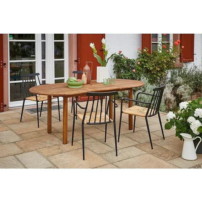 Envy Gartensessel Eva - Rattan/Geflecht, Stapelbar, Schwarz/Natur, B x T x H: 58 x 54 x 84 cm - Das Bild zeigt einen ovalen Holztisch mit vier schwarzen Stühlen mit Rattan-Sitzen auf einer Terrasse, umgeben von Pflanzen und Blumen.