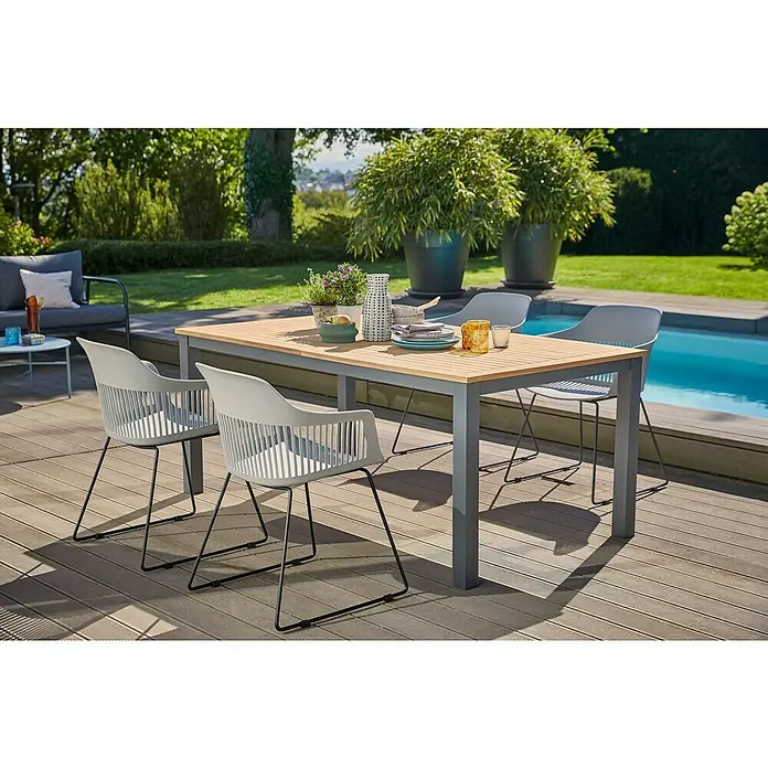 Sunfun Gartenmöbel-Set Carina/Annika - 5 -tlg., Holz, Naturbraun/Anthrazit/Hellgrau, Tischplatte ausziehbar - Ein einladender Esstisch mit vier Stühlen auf einer Terrasse, ideal für Mahlzeiten im Freien mit Blick auf den Pool und die grüne Umgebung.