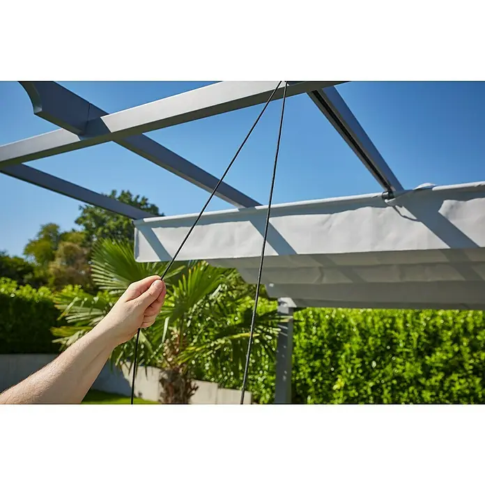 Sunfun Raffpavillon Kreta - B x T: 418 x 333 cm, Farbe Dach: Hellgrau - Detailaufnahme einer Person, die an der Zugschnur einer Pergola zieht, um das Sonnendach zu bedienen, im Hintergrund grüne Vegetation.