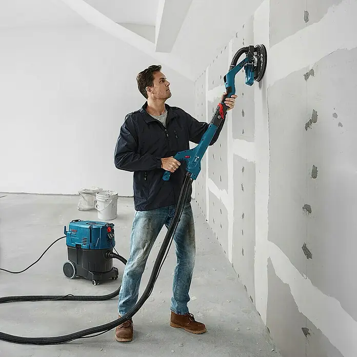 Bosch Professional Lijadora de pared y techo GTR55-225 - 550 W, Diámetro disco de lija: 225 mm - Un hombre utilizando una lijadora de paneles de yeso de Bosch en una pared, conectada a una aspiradora para un lijado sin polvo.