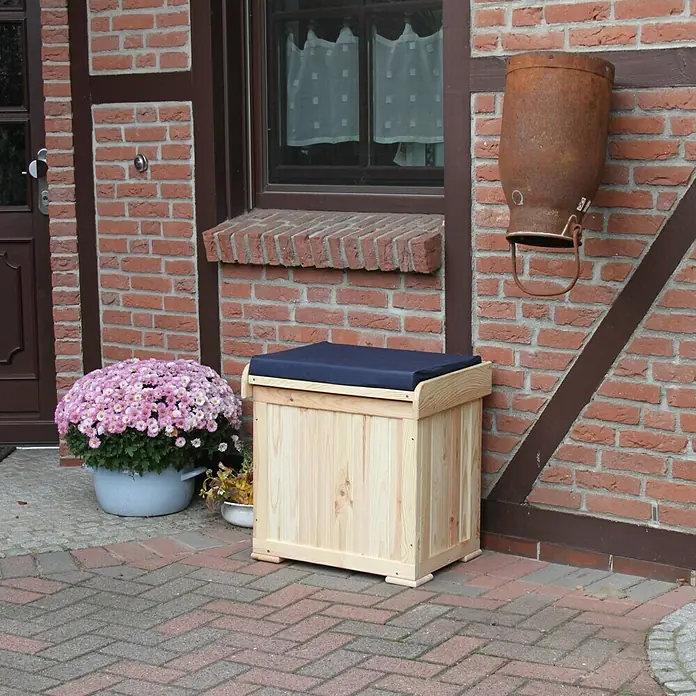 Promadino Auflagenbox Patrick - 56 x 43 x 56 cm, Holz, Natur, Mit Auflage, Mit Sitzfunktion - Eine hellholzfarbene Sitzbank mit dunkelblauem Kissen steht vor einer Backsteinmauer und Blumenkübeln.