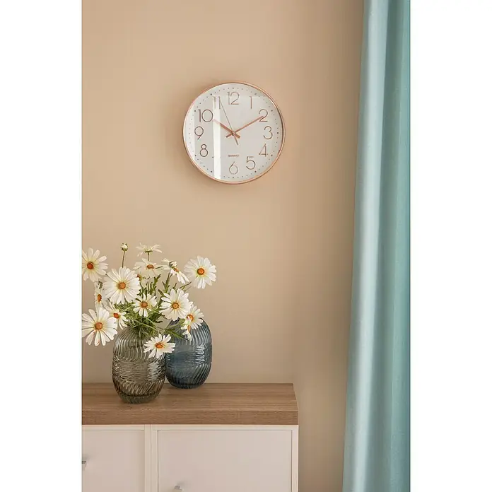 Wanduhr Lille - Durchmesser: 30 cm, Weiß/Rosé - Eine Nahaufnahme einer dekorativen Wanduhr mit römischen Ziffern, die über einer Kommode mit Blumen und Vasen hängt.