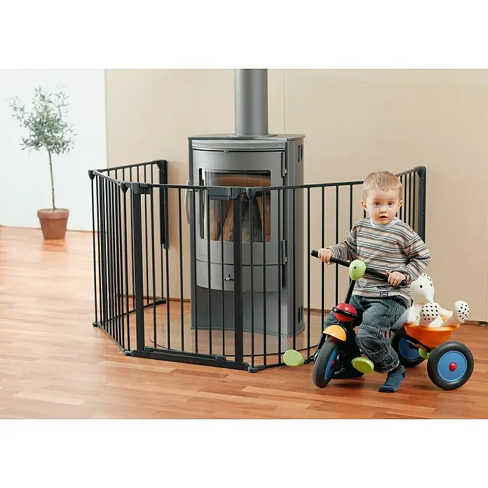 Star Stairs Sicherheitsgitter Ben - L x H: 278 x 71 cm, Geeignet für: Kinder bis 24 Monate - Ein Kind auf einem Dreirad vor einem schwarzen Ofen, der mit einem Sicherheitsgitter umgeben ist.