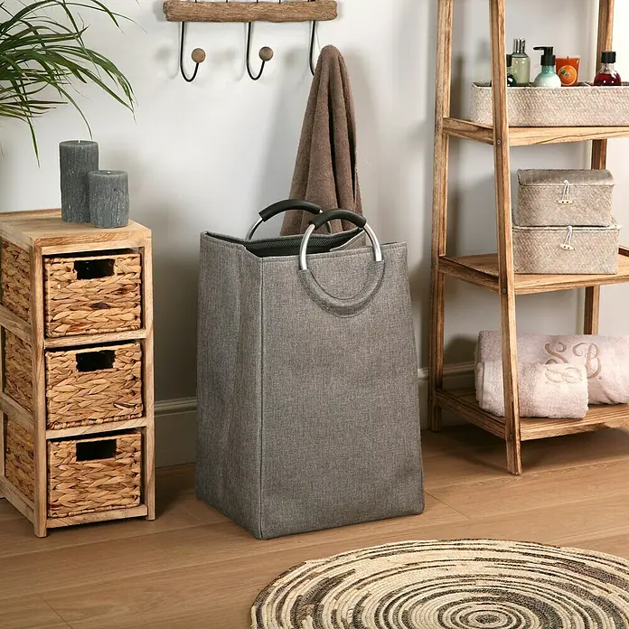 Versa Cesta para la ropa con asas - L x An x Al: 46 x 30 x 55 cm, Gris - Un cesto de ropa sucia gris con asas redondas está en un baño, al lado de un armario tejido y una estantería de madera.