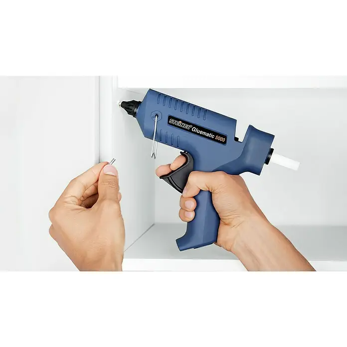 Steinel Klebepatronen Weiß - 10 Stk., Weiß, Durchmesser Klebepatrone: 11 mm - Eine blaue Steinel Gluematic 5000 Klebepistole wird mit einer Hand bedient, während die andere einen Klebestift hält.