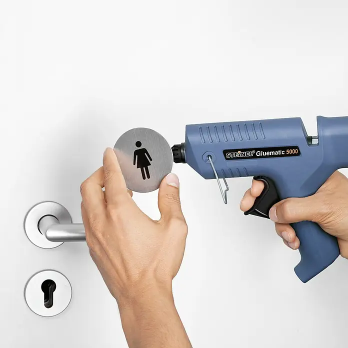 Steinel Heißklebepistole Gluematic 5000 - Durchmesser Klebepatrone: 11 mm, 500 W - Das Bild zeigt eine Steinel Gluematic 5000 Heißklebepistole, die ein Frauensymbol an einer Tür befestigt. Praktisches Werkzeug für den täglichen Gebrauch.
