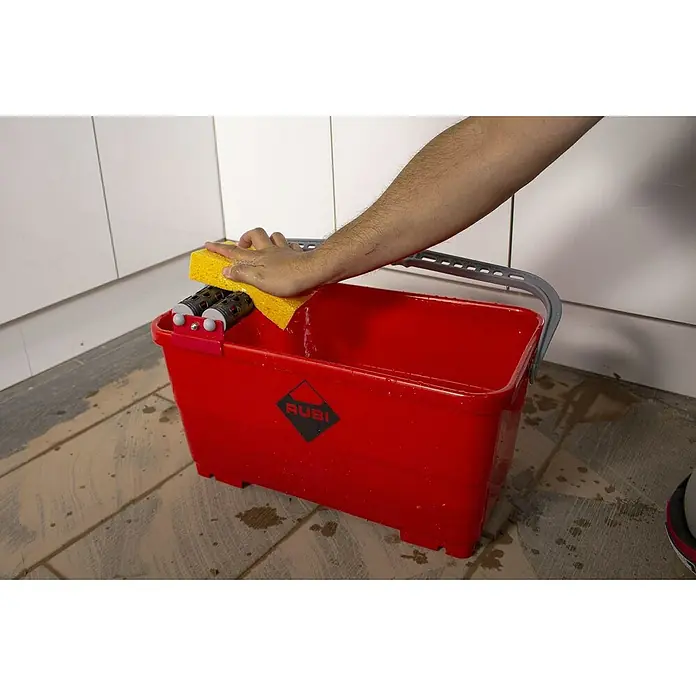 Rubi Cubeta de limpieza Easypro - Polipropileno, 24 l - Un primer plano de un cubo rojo Rubi con rodillos escurridores para trabajos de azulejos, que está siendo limpiado por una mano con una esponja amarilla.