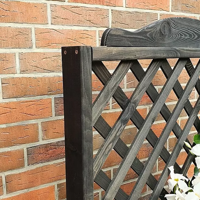Promadino Pflanzkasten Romantica - Außenmaß (L x B x H): 80 x 38 x 140 cm, Holz, Anthrazit - Dunkles Holzspalier mit diagonalen Leisten, geschmückt mit weißen Blumen, vor einer rotbraunen Ziegelwand.